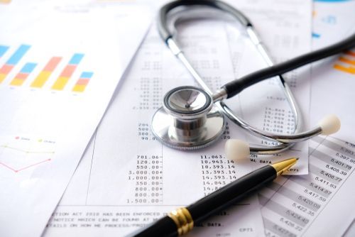 A stethoscope and a black pen rest on top of medical financial documents and a colorful bar chart.