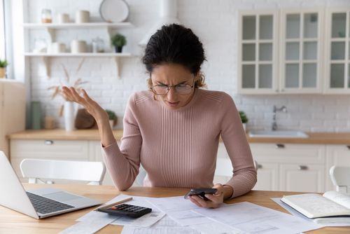 A person in a light pink sweater looks frustrated while checking a phone and calculator at a desk with paperwork.