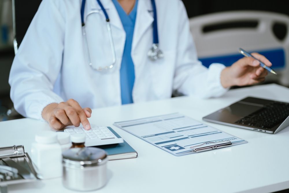 A healthcare professional in a white coat sits at a desk with a medical chart, calculator, and laptop.