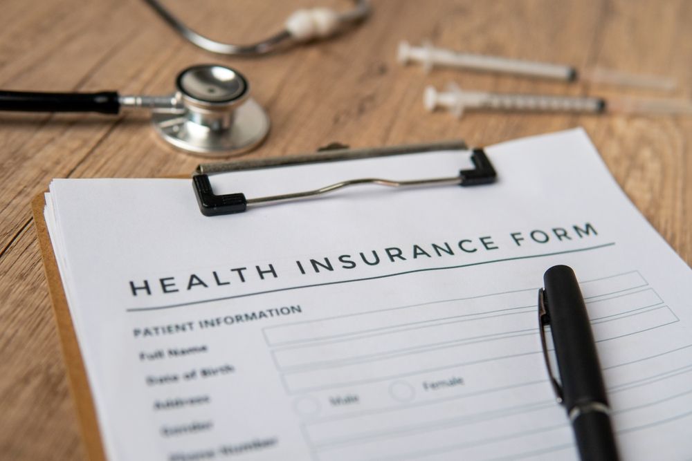 A health insurance form on a clipboard with a stethoscope and syringes nearby on a wooden desk.