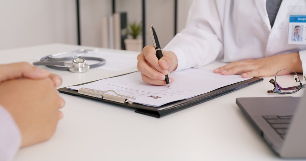 A doctor in a white coat writes on a clipboard during a consultation with a patient at a desk with a stethoscope.