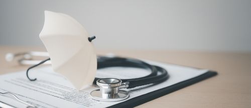 A stethoscope rests on a medical document with a small white umbrella placed on top, symbolizing health insurance.