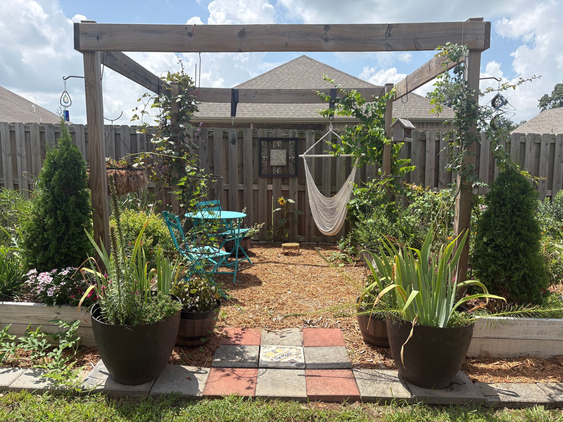 Backyard patio garden with wooden pergola, hanging chair, and potted plants leading to a fenced gate.