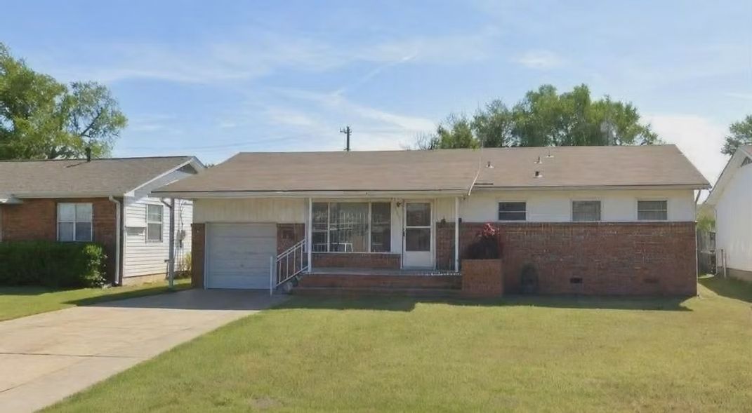Single-story brick house with a front porch, garage, and a green lawn under a clear sky