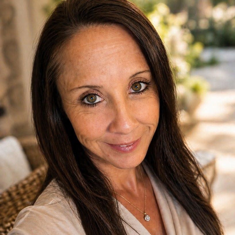Close-up portrait of a smiling woman in a beige top with long dark hair outdoors.