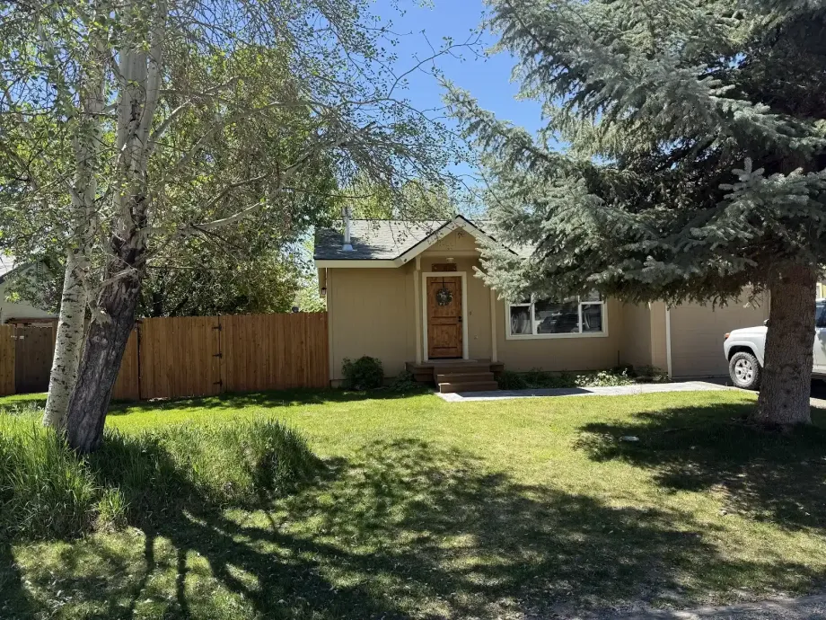 Small beige house with a front lawn, wooden fence, and trees in the yard.