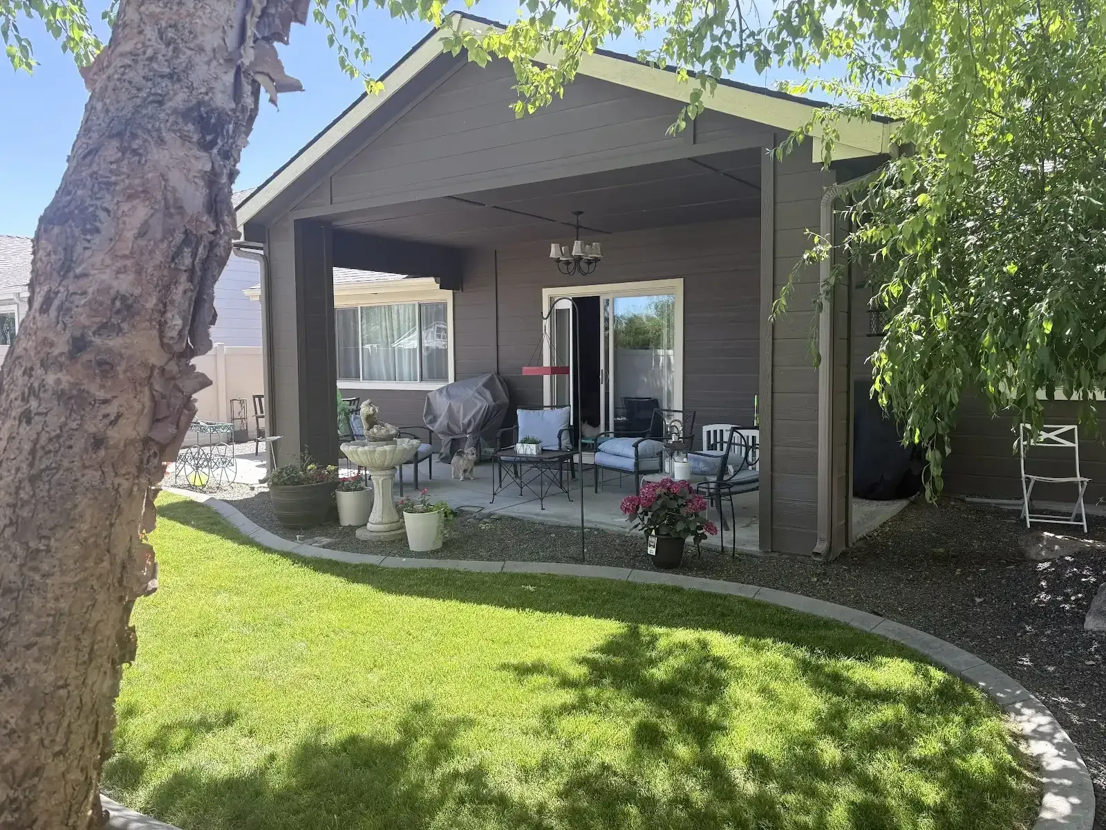 Covered patio with seating, potted plants, and a green lawn beside a house