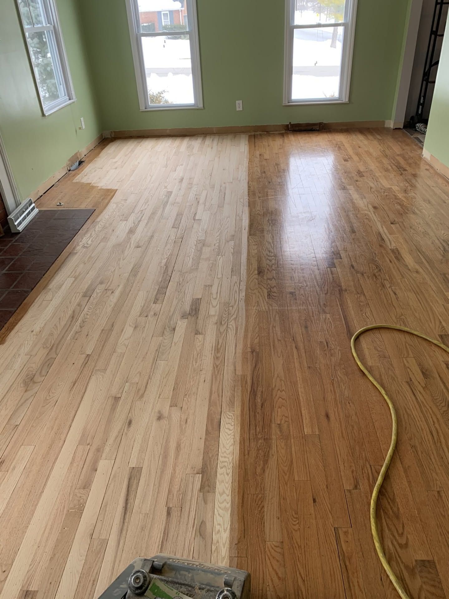 Partially sanded hardwood floor in an empty room with green walls and windows; sander and hose visible