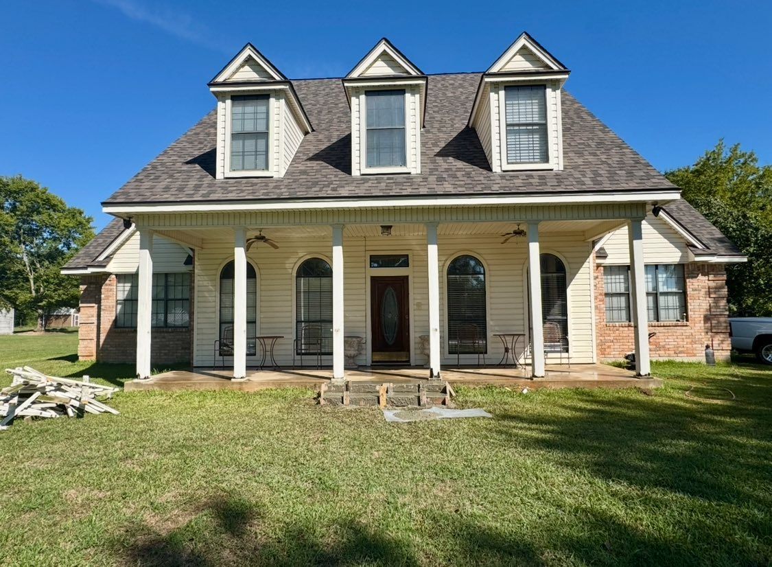 A two-story, cream-colored house with a large front porch, three dormer windows on the roof, and a grassy front yard.