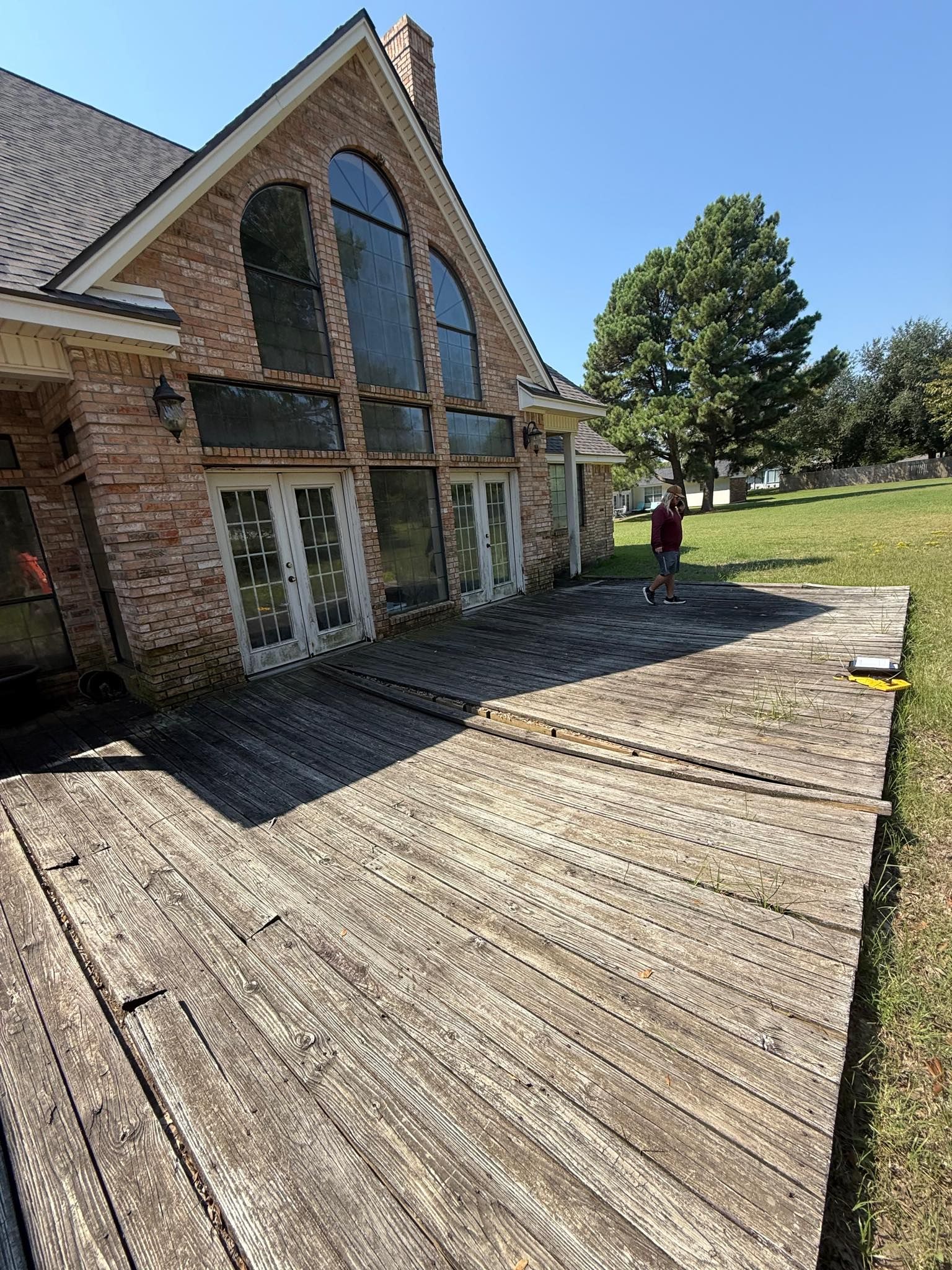 A weathered, dark wooden deck extends from a brick house with large windows under a clear blue sky.