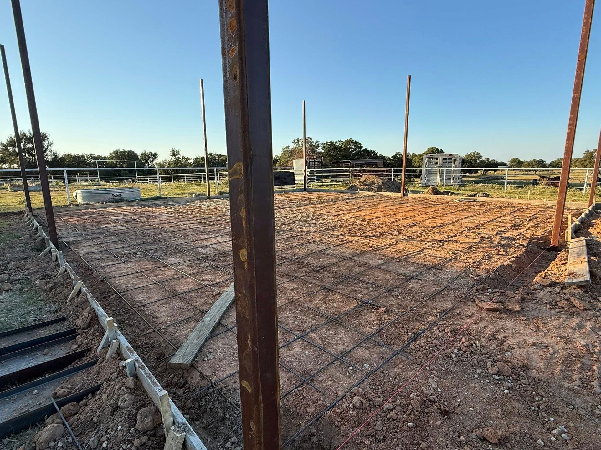 Construction site showing a dirt foundation prepped with metal wire mesh grid and metal posts against a clear blue sky.