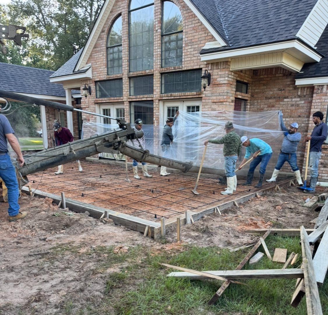 Construction workers using a concrete pump truck to pour a foundation in front of a brick house.