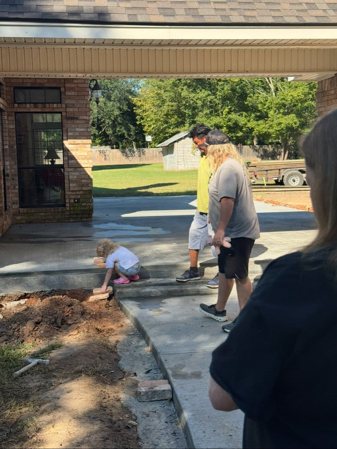 A child plays on a concrete patio while two people stand nearby, looking on during an outdoor construction project.