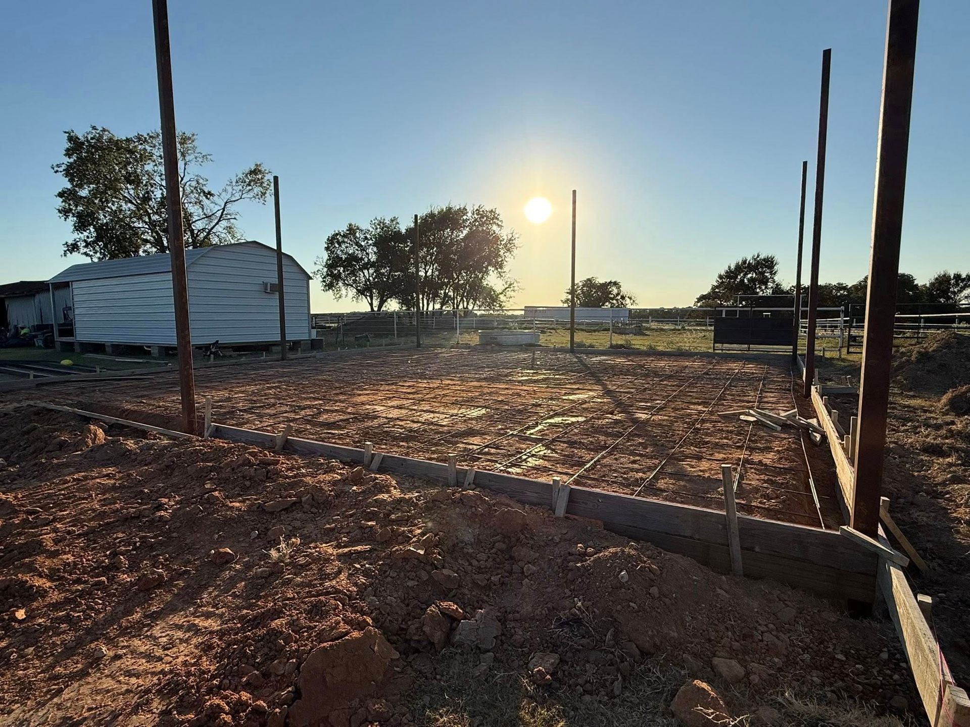 Construction site at sunset with metal poles, wooden formwork boards, and cleared dirt ground under a clear blue sky.
