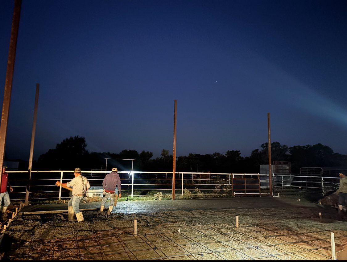 Two workers spread wet concrete across an outdoor construction site at twilight, framed by metal poles and fencing.