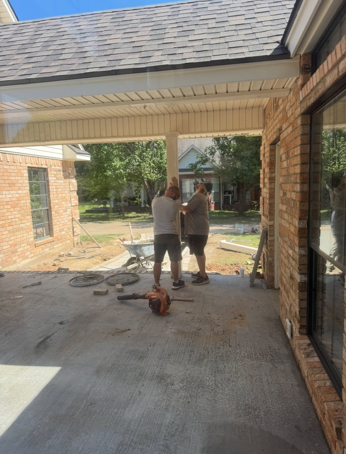Two people work on a project on a concrete patio outside a brick house.