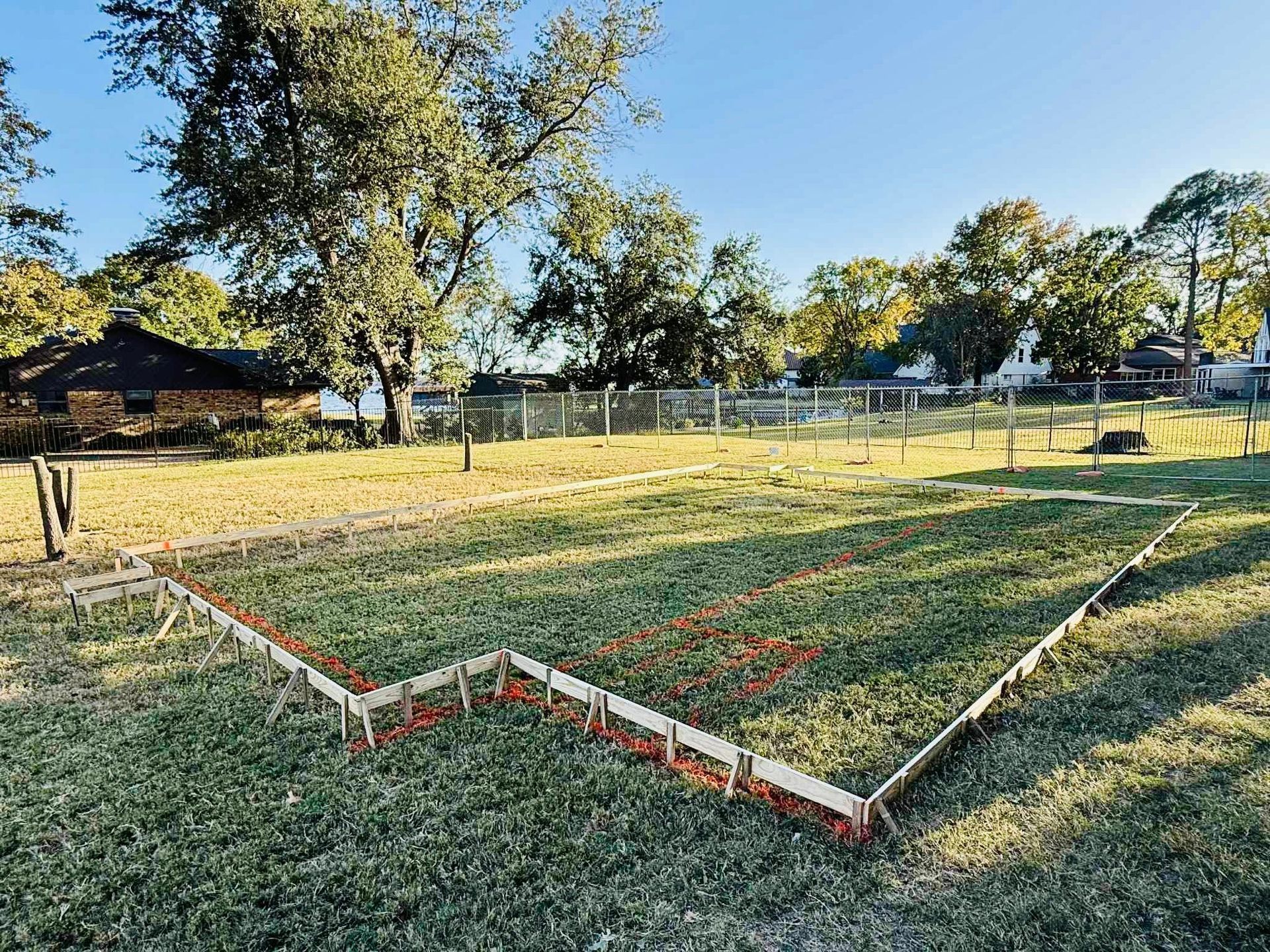 A grassy yard features a wooden form laid out on the ground, outlined with red spray paint to prepare for a new foundation.