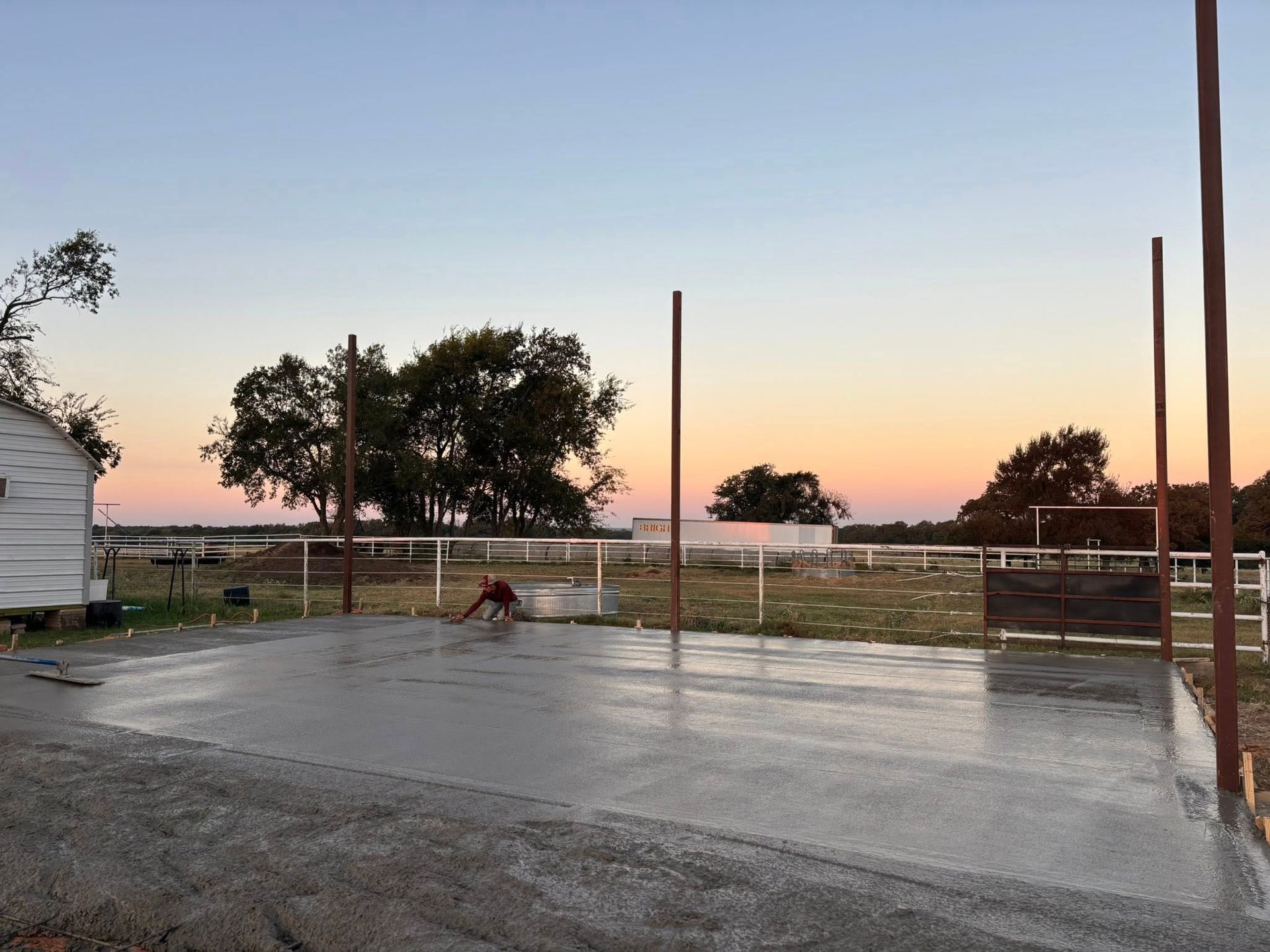 A person finishes a wet concrete slab outdoors at sunset, framed by metal poles and a rural landscape.