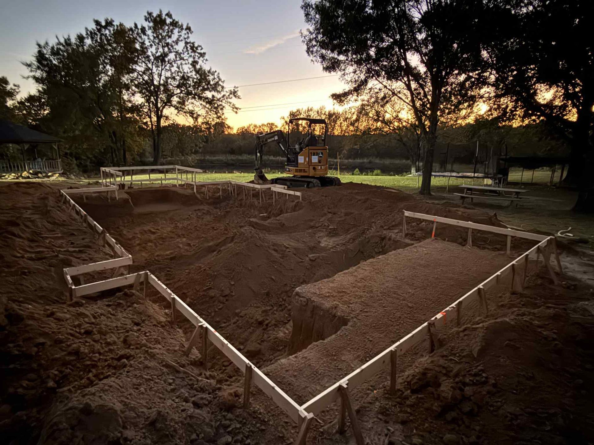 A construction site at sunset showing a deep pool excavation with wooden formwork in a rural backyard.