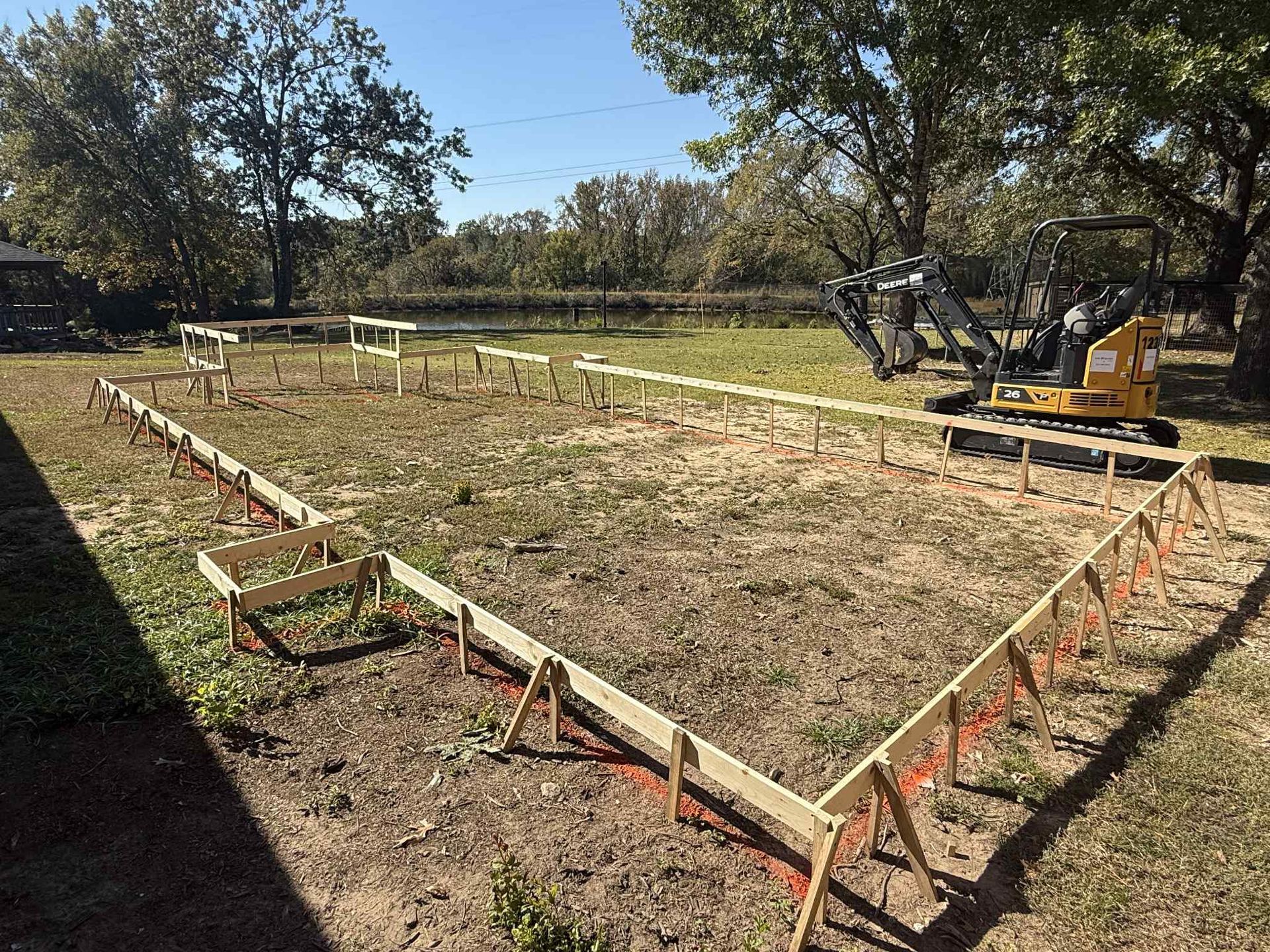 A yellow excavator sits by a wooden construction frame outlining a rectangular building foundation on a grassy lot.
