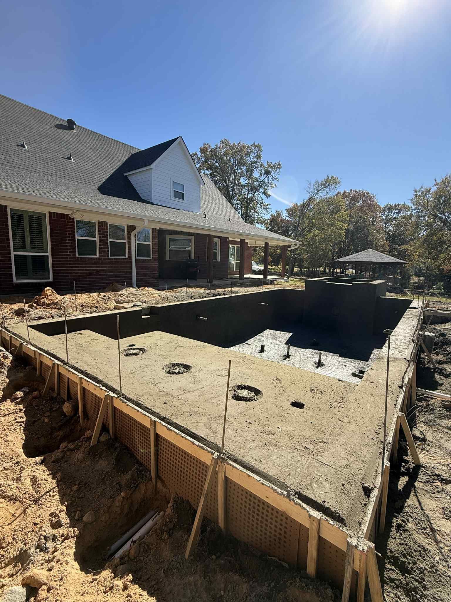 A partially constructed swimming pool foundation with wooden framing in front of a brick house under a bright sunny sky.