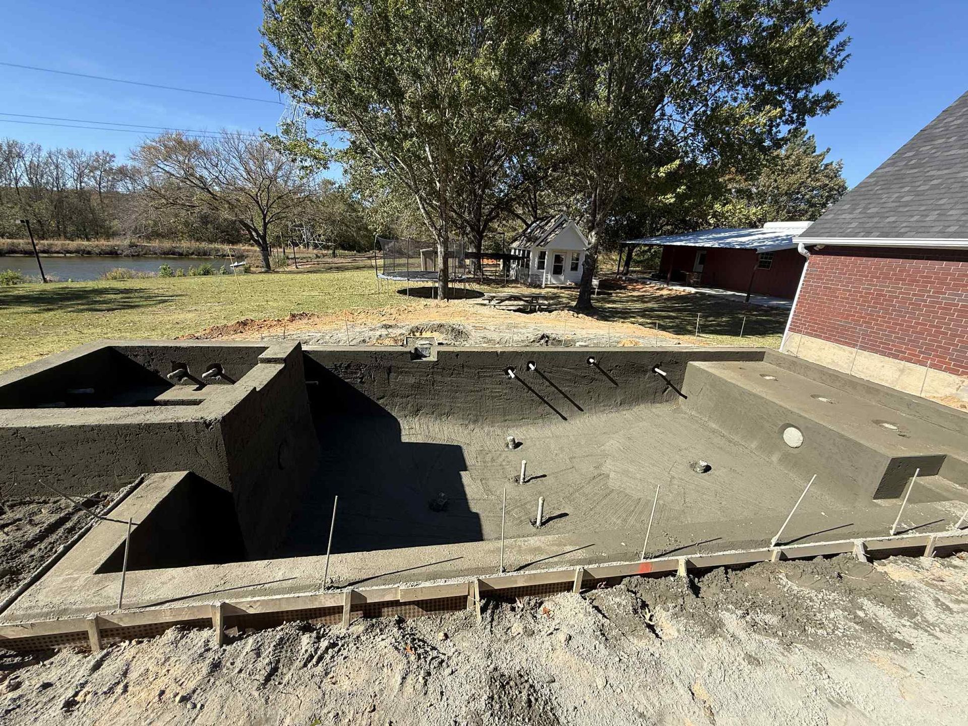 An unfinished concrete swimming pool under construction in a grassy yard, next to a brick building.
