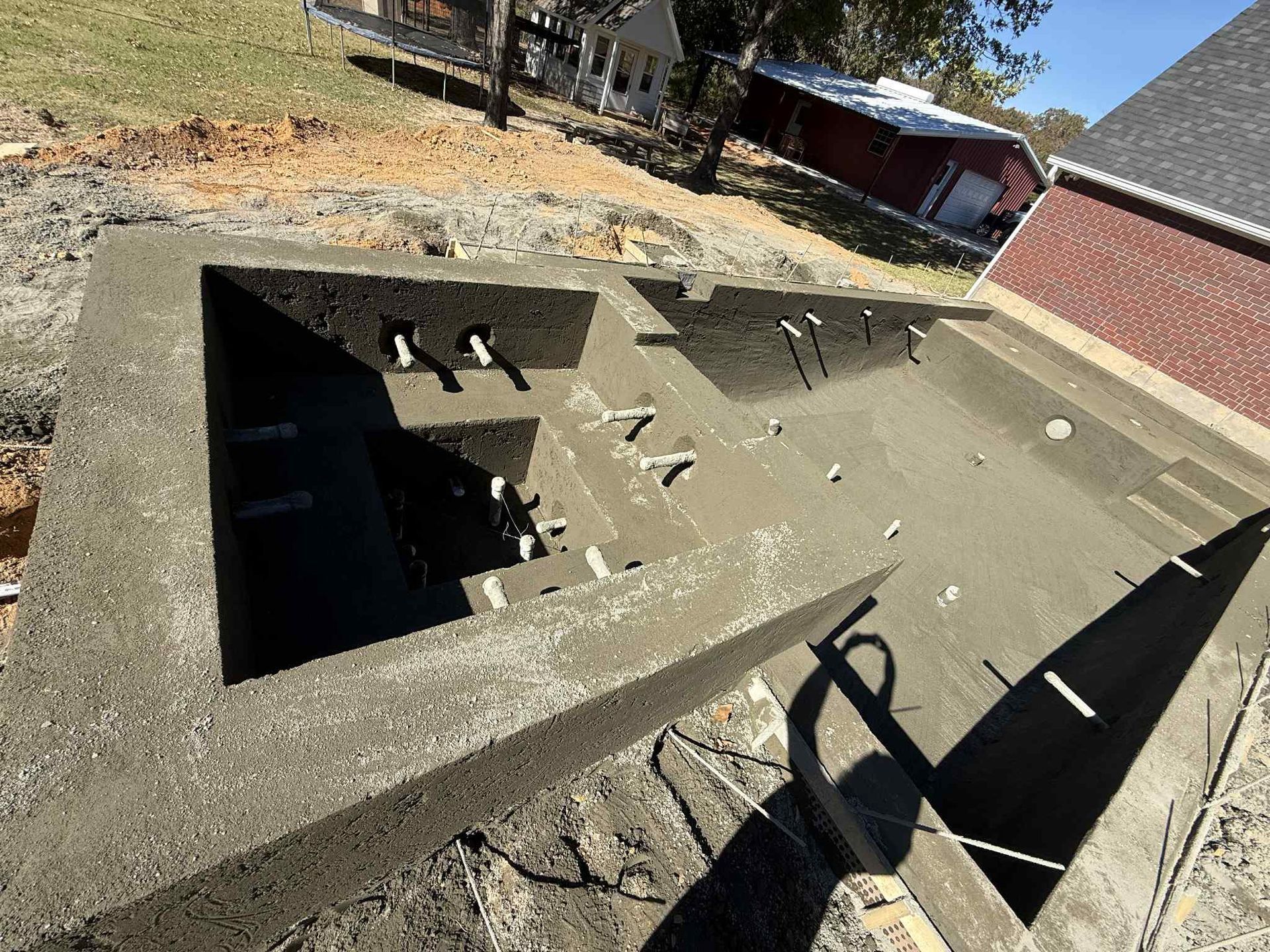 An unfinished swimming pool with concrete walls and exposed plumbing pipes in a residential backyard.