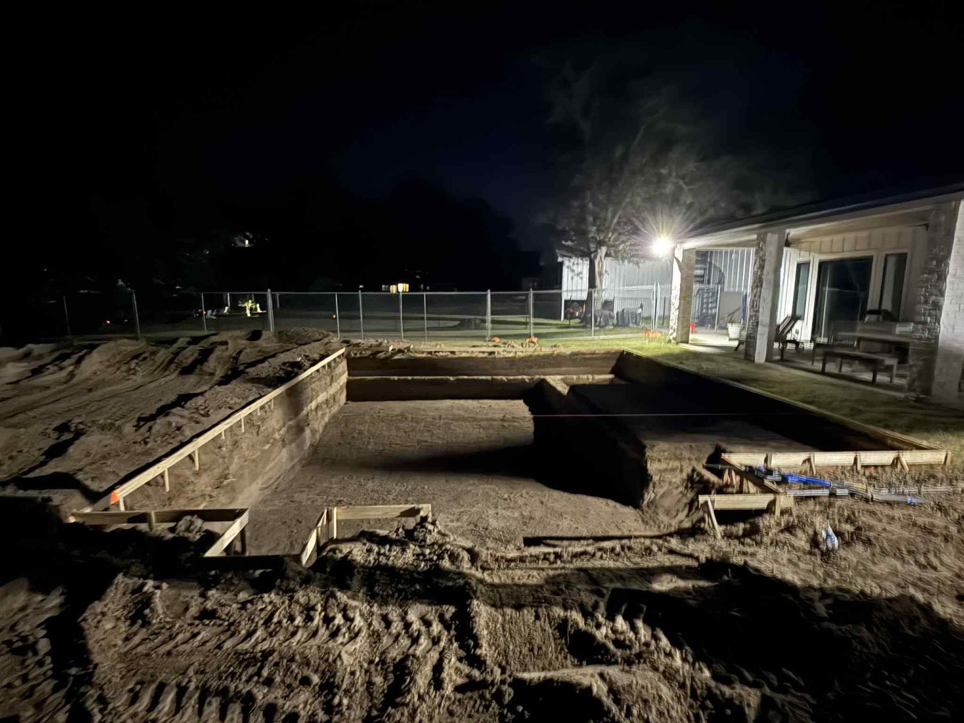 Night view of a backyard swimming pool under construction, showing a deep excavated pit with wooden forms and soil.