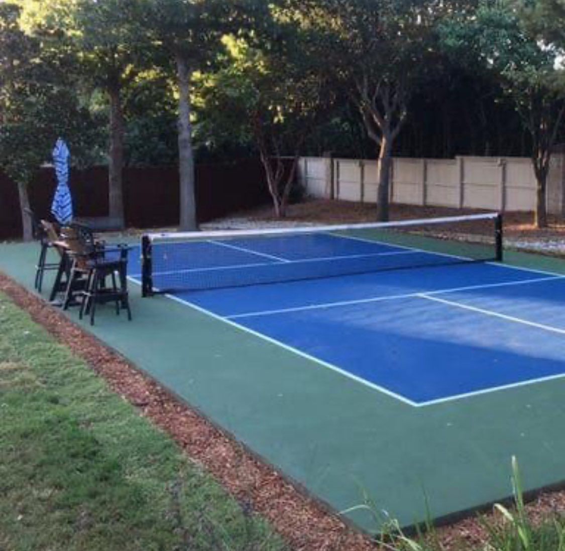 A blue and green pickleball court outdoors, surrounded by trees, with chairs and a blue umbrella on the side.