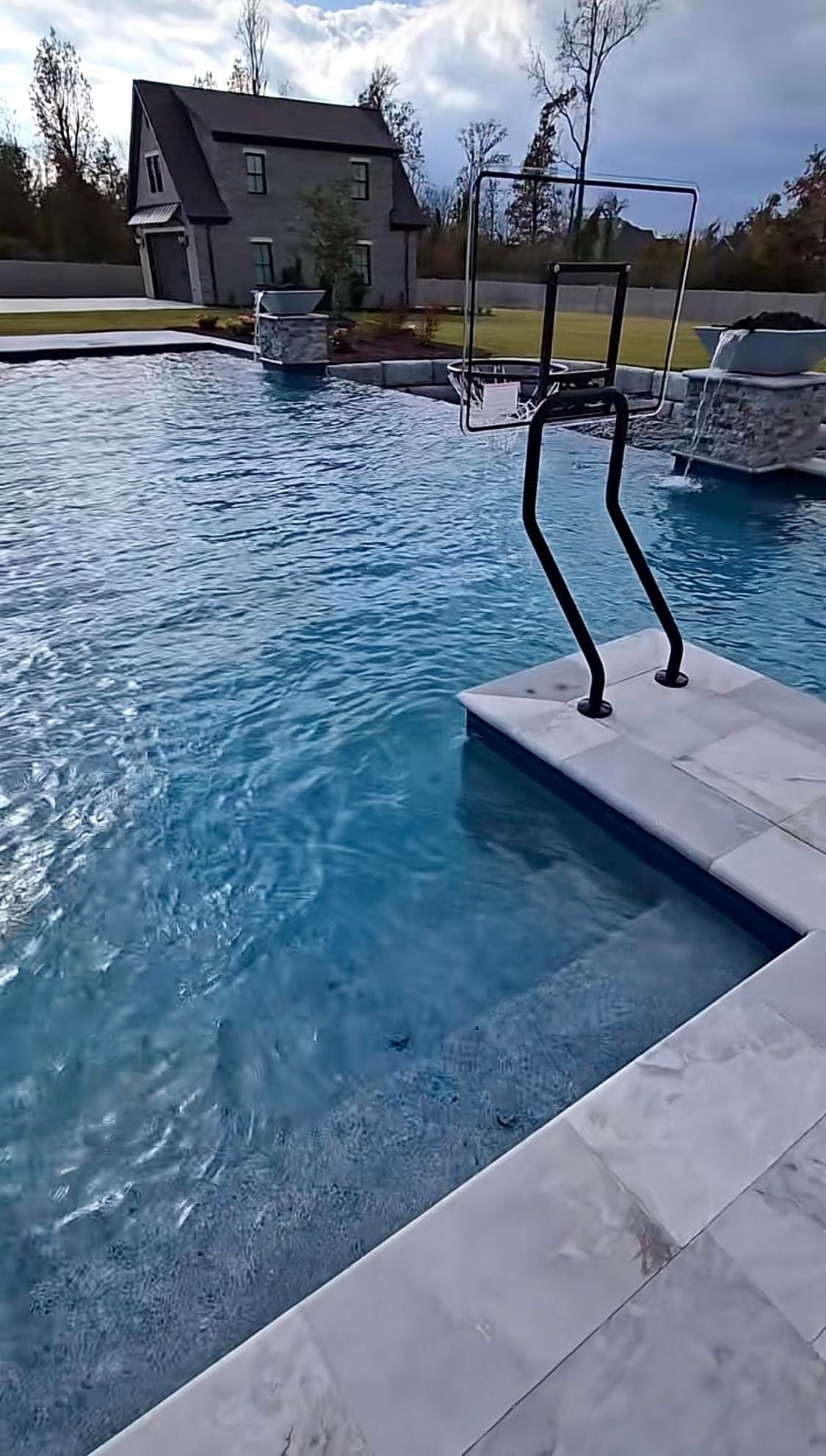 A swimming pool with blue water and a basketball hoop near the edge, set in a backyard with a house in the background.