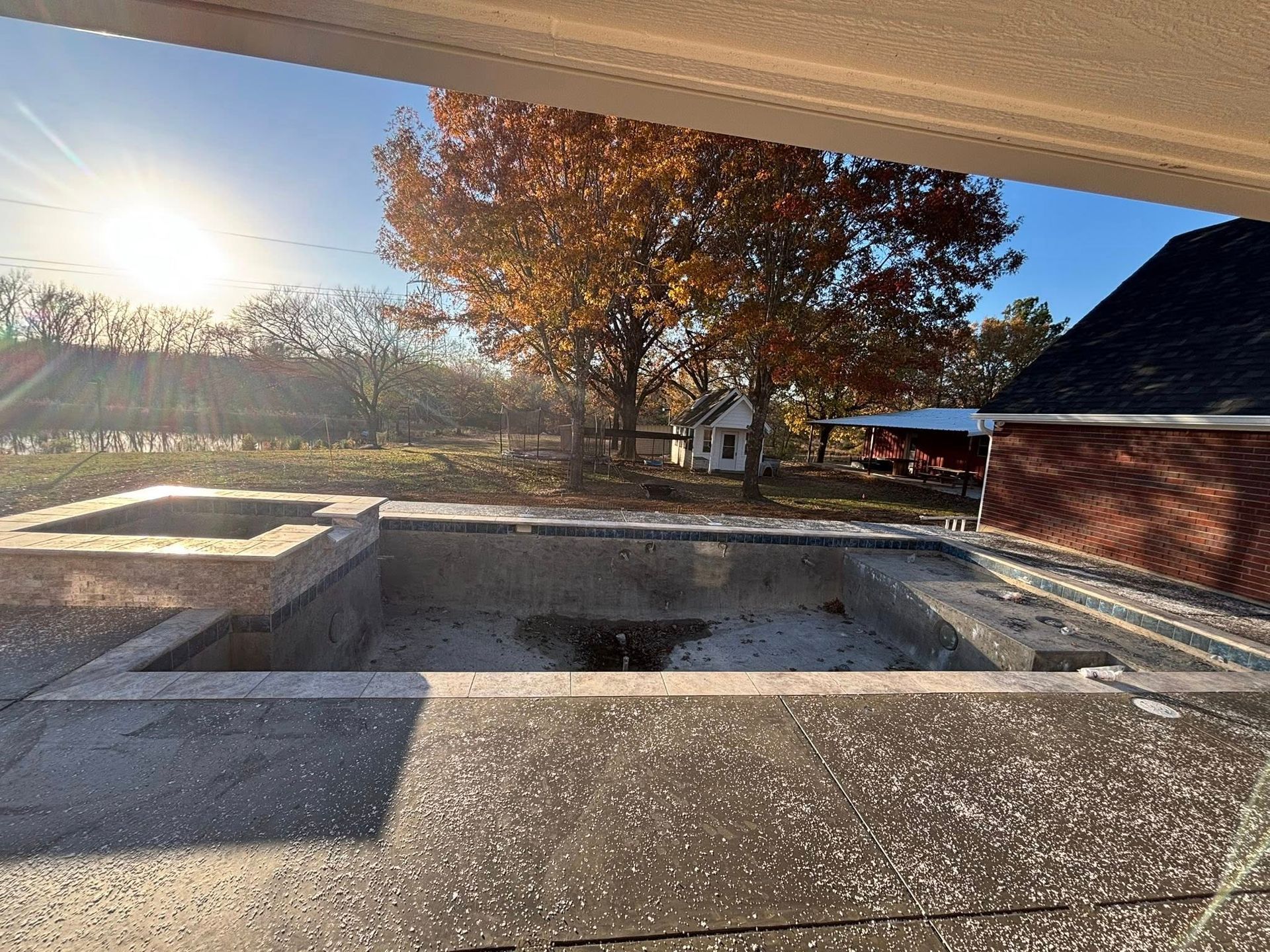 An empty concrete pool with a small brick fireplace on a sunny day with fall trees in the background.