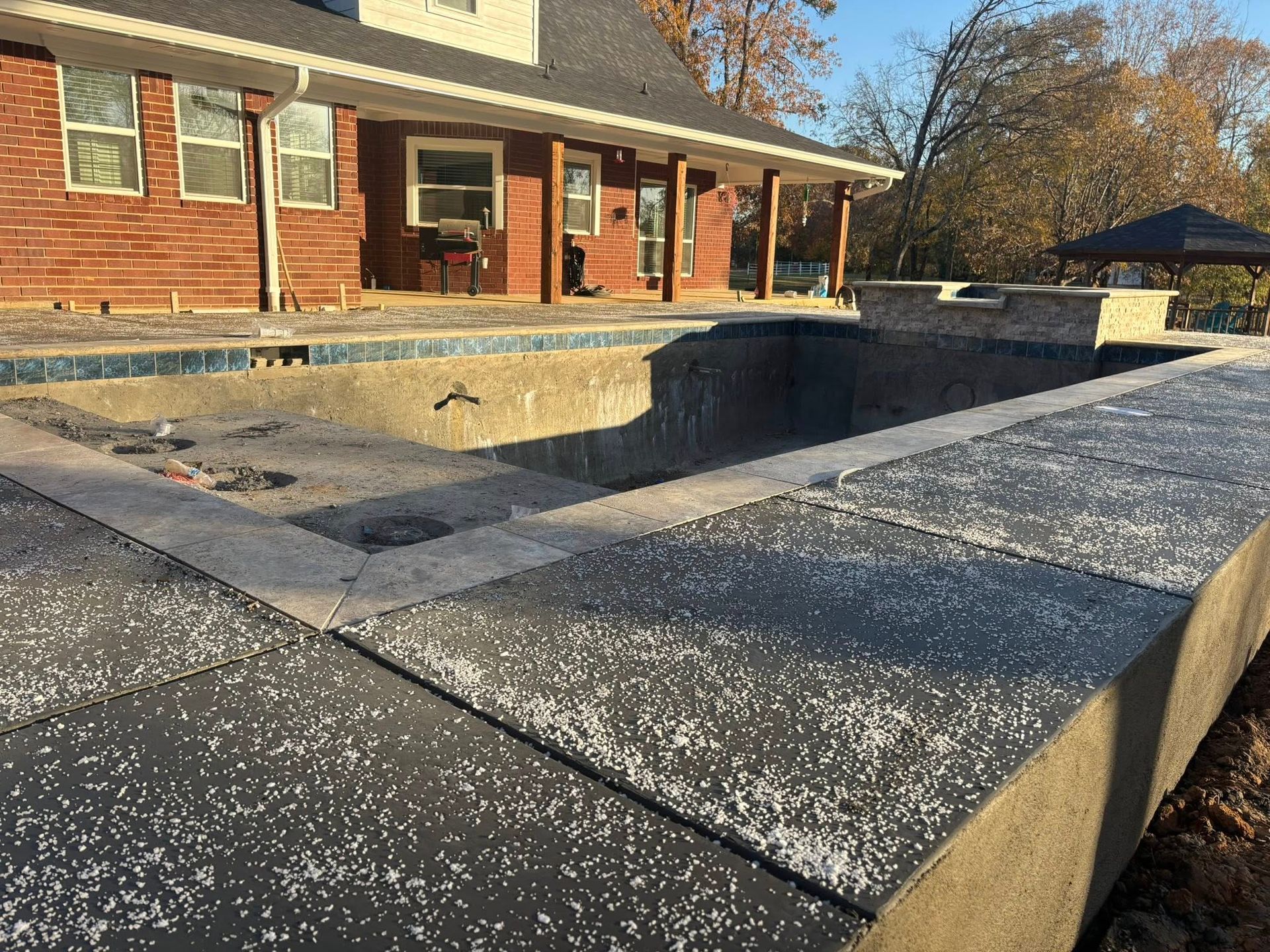 A partially constructed swimming pool and patio area next to a brick house on a sunny day.