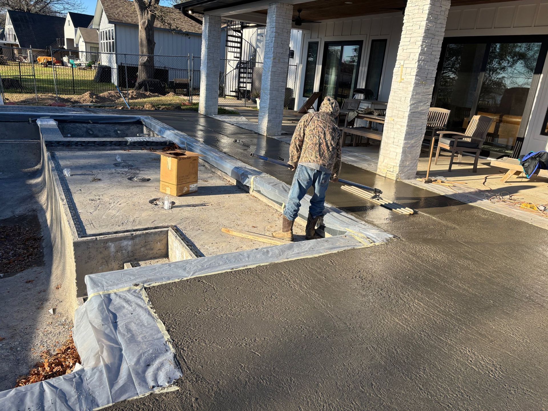 A person in a hooded jacket walks on a newly poured concrete patio beside an unfinished pool frame.