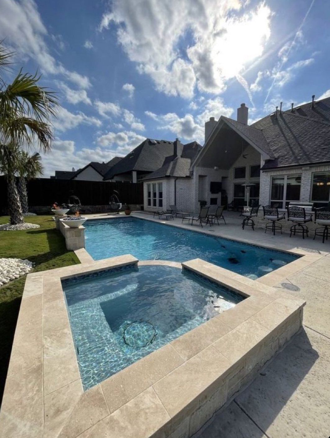 A rectangular swimming pool and raised square spa with light stone decking in a sunny backyard with a large house behind.