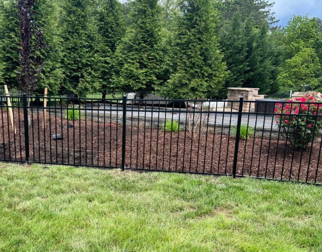 A black metal fence separates a green lawn from a garden bed, evergreen trees, and a stone outdoor fireplace.