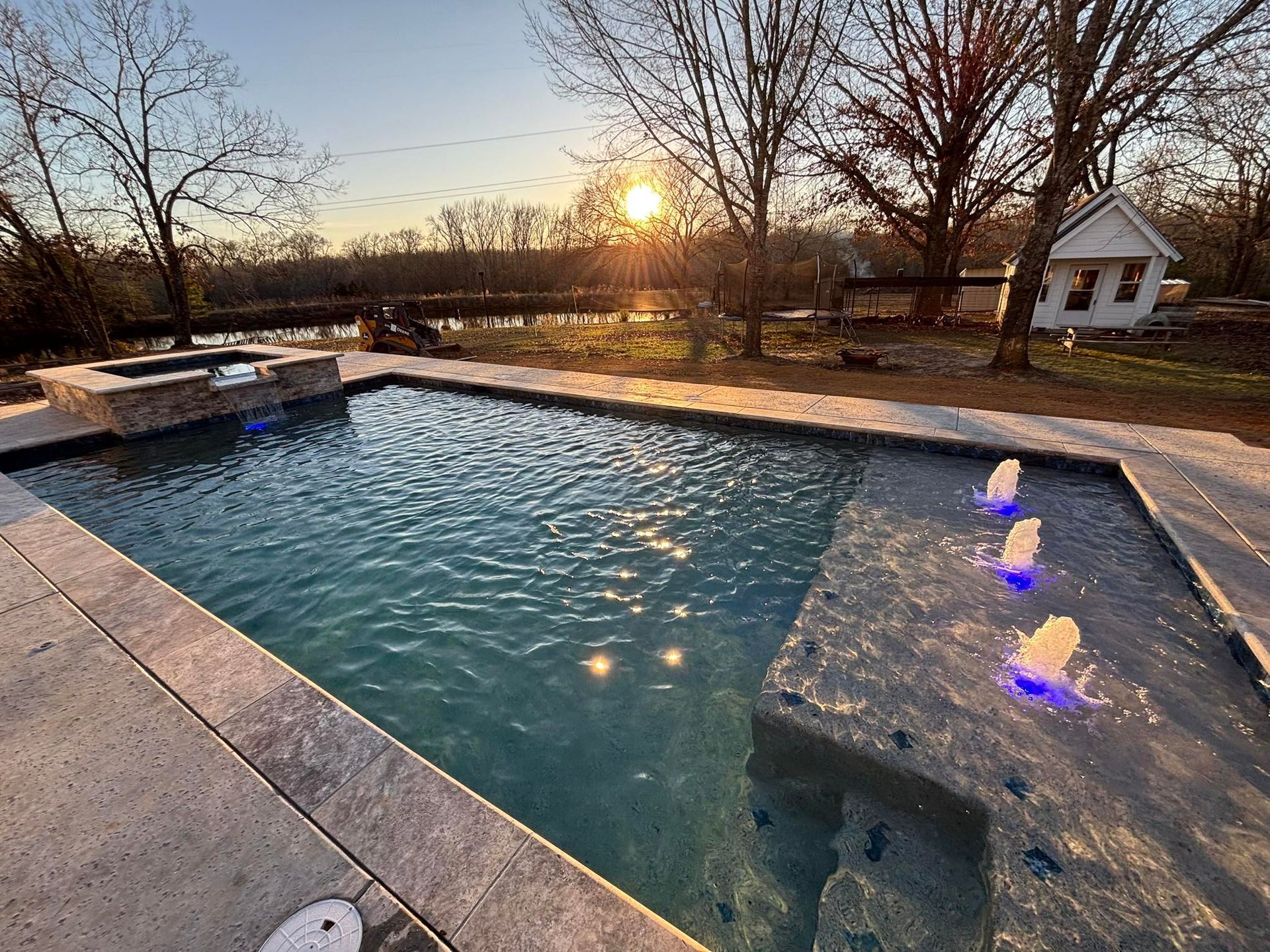 A swimming pool with glowing water jets and a stone hot tub at sunset, set in a backyard near trees and a small building.