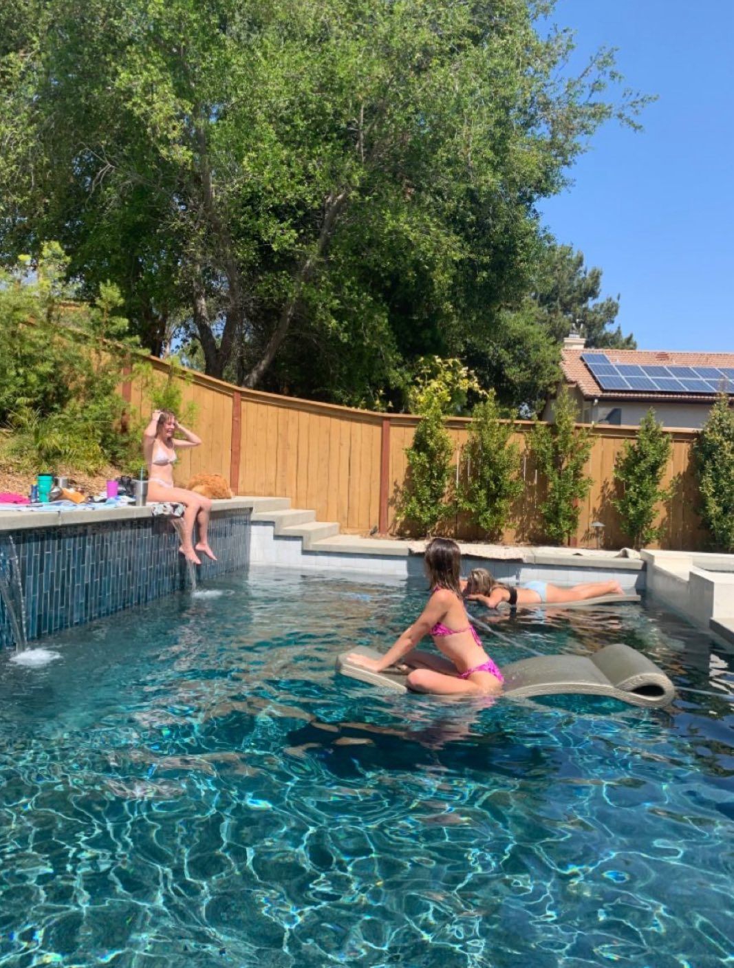 Three people enjoying a sunny day in a swimming pool, with two sitting on loungers and one relaxing on the pool edge.