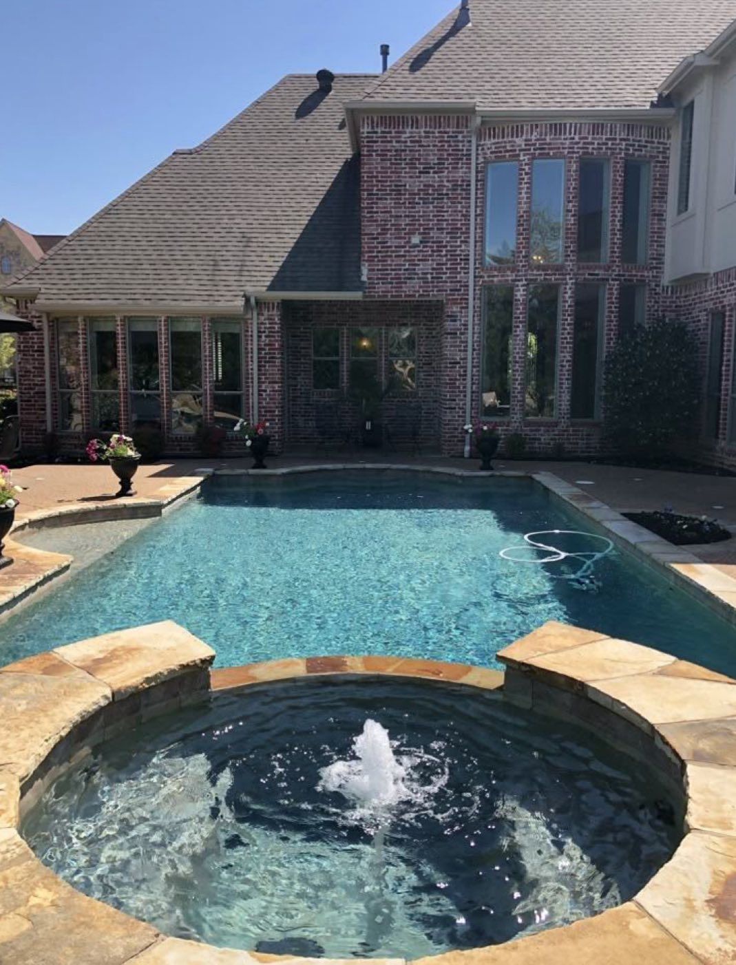 A backyard swimming pool and circular hot tub with a fountain in front of a multi-story red brick house.