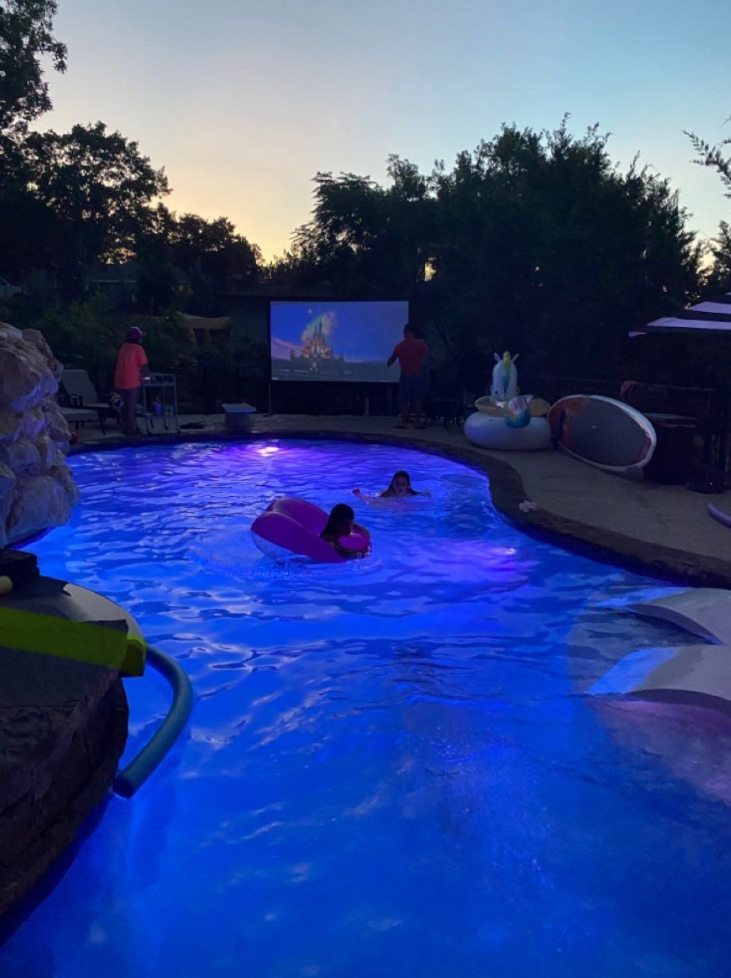 People swim in a blue-lit pool at dusk while watching a movie on an outdoor screen.