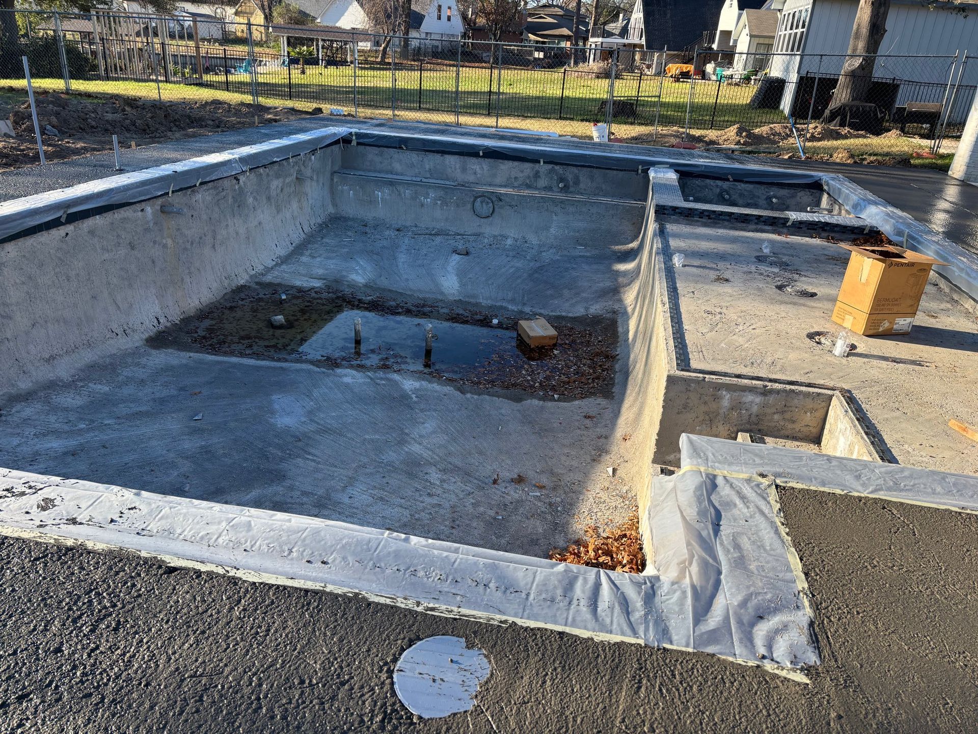 An empty rectangular concrete swimming pool under construction in a backyard, with a small pool of water in the center.