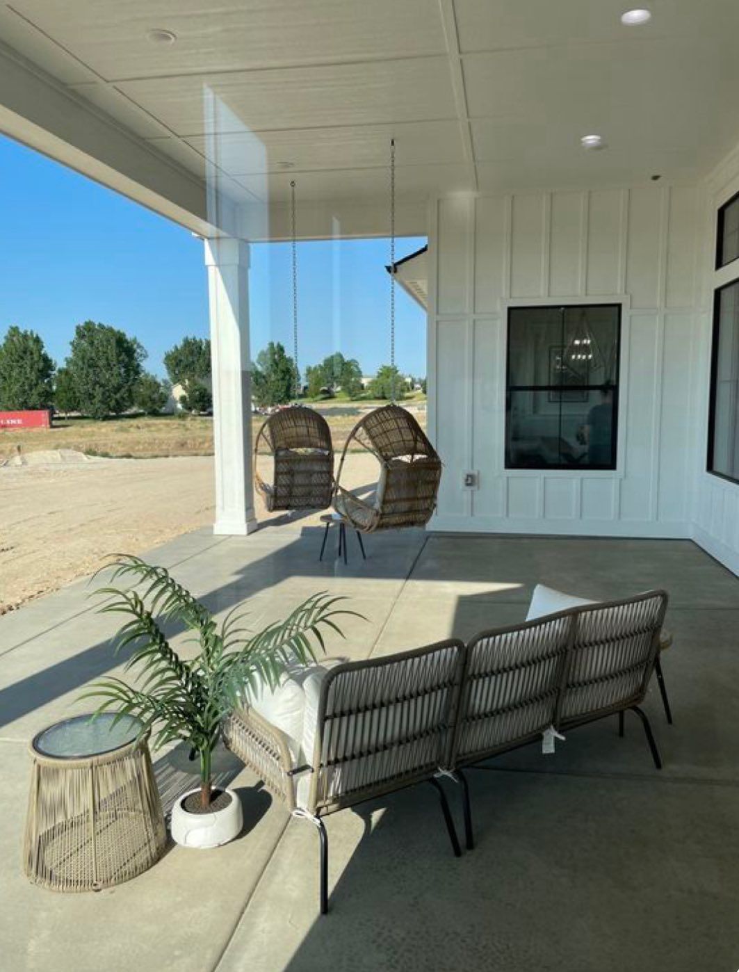 A covered patio with a woven outdoor sofa, two hanging egg chairs, and a small side table featuring a potted plant.