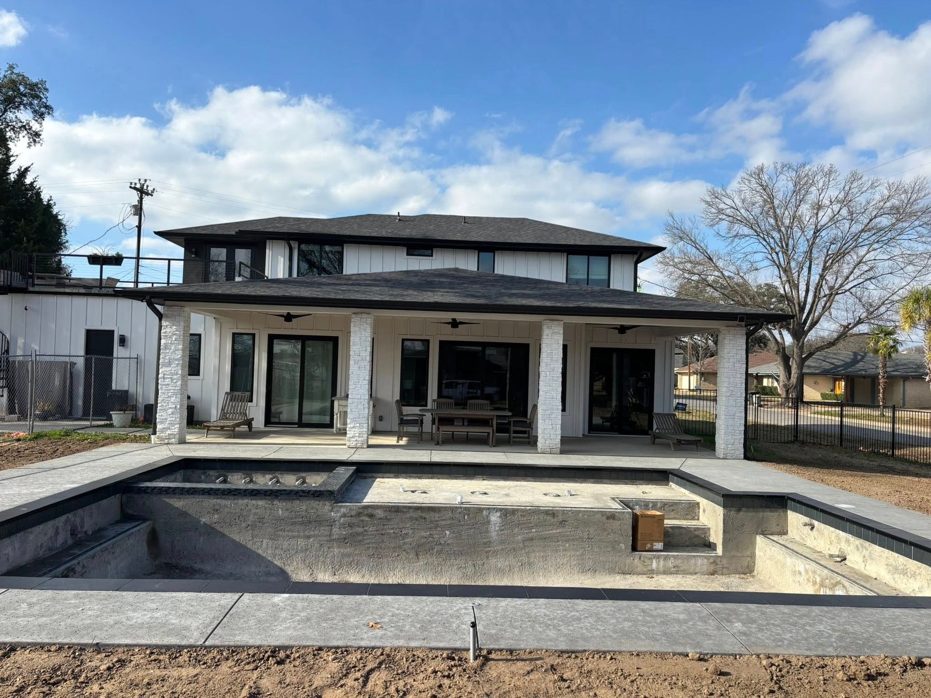 A two-story house with a covered patio overlooks an empty, unfinished rectangular swimming pool under a blue sky.