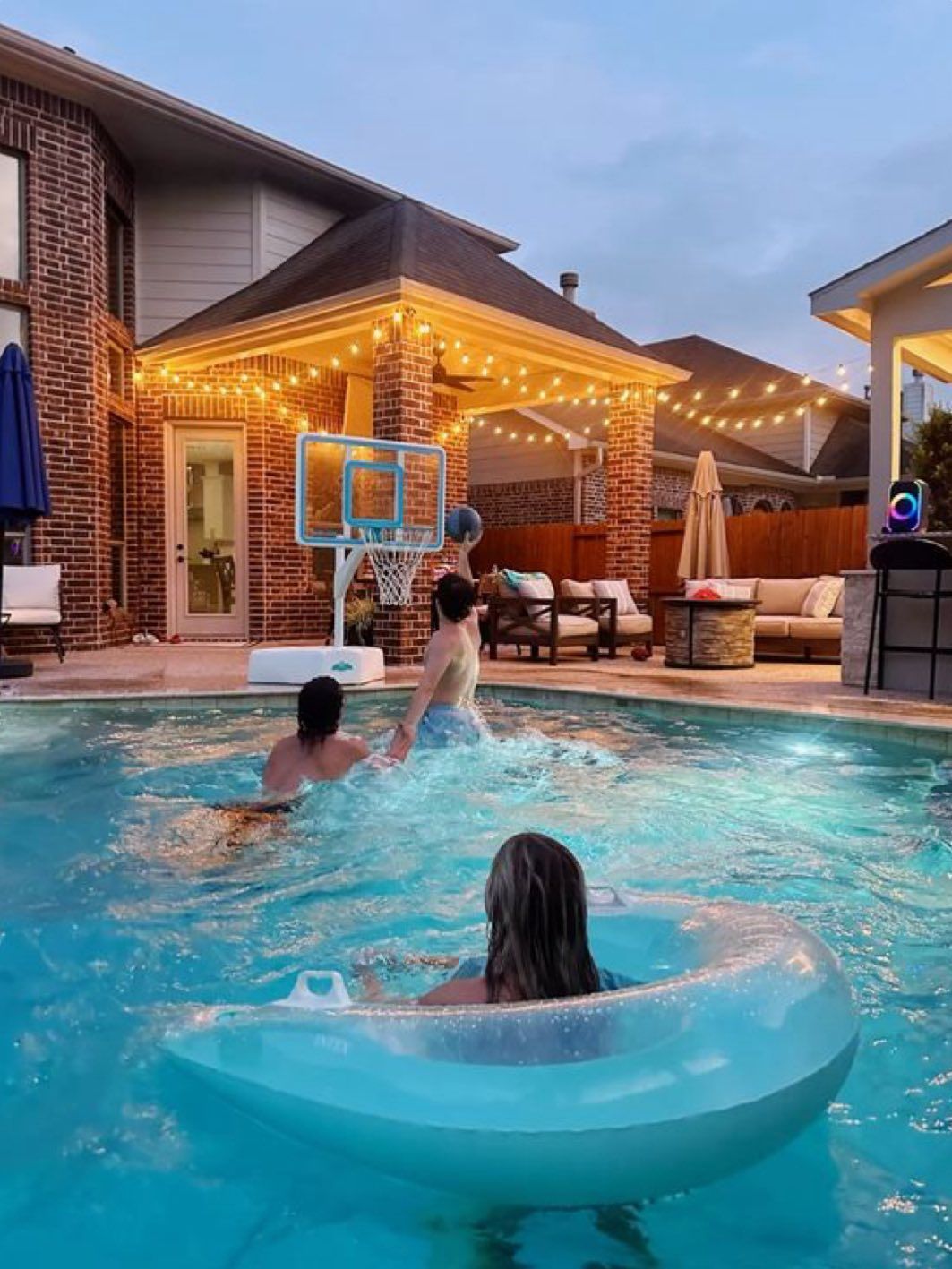 People swimming and playing basketball in a backyard pool at twilight, lit by string lights over a patio.