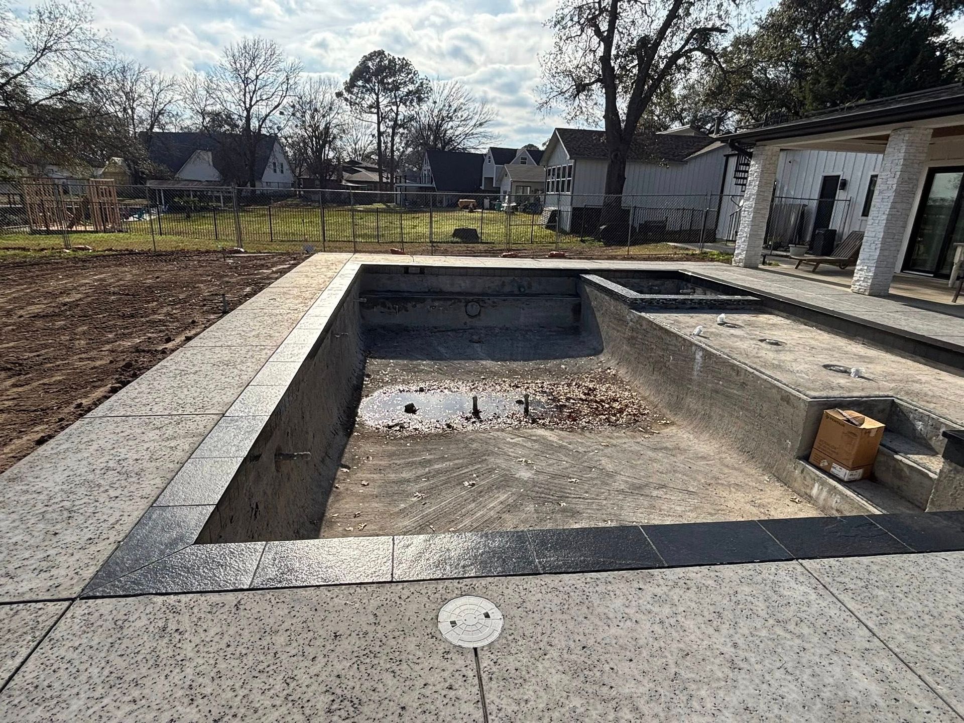 An unfinished backyard swimming pool with dark coping and light-colored concrete decking under a bright sky.