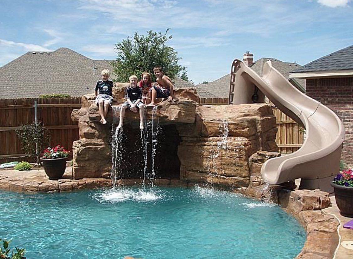 Four children sit atop a backyard swimming pool rock formation with a waterfall and a curved slide.