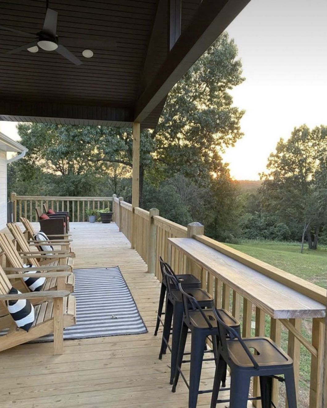 A wooden deck with Adirondack chairs, a striped rug, and a bar-style railing with black stools overlooking a forest.