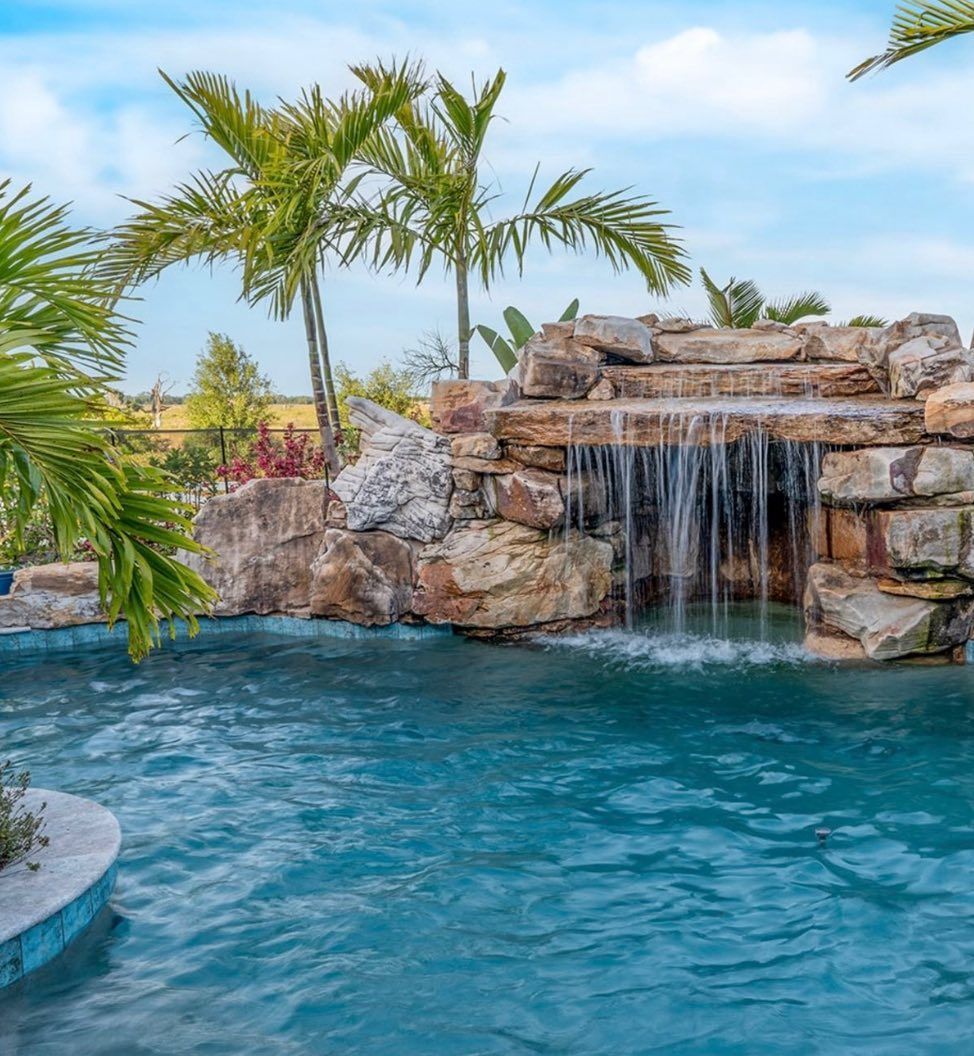 A rocky stone waterfall flows into a blue backyard swimming pool surrounded by palm trees under a bright blue sky.