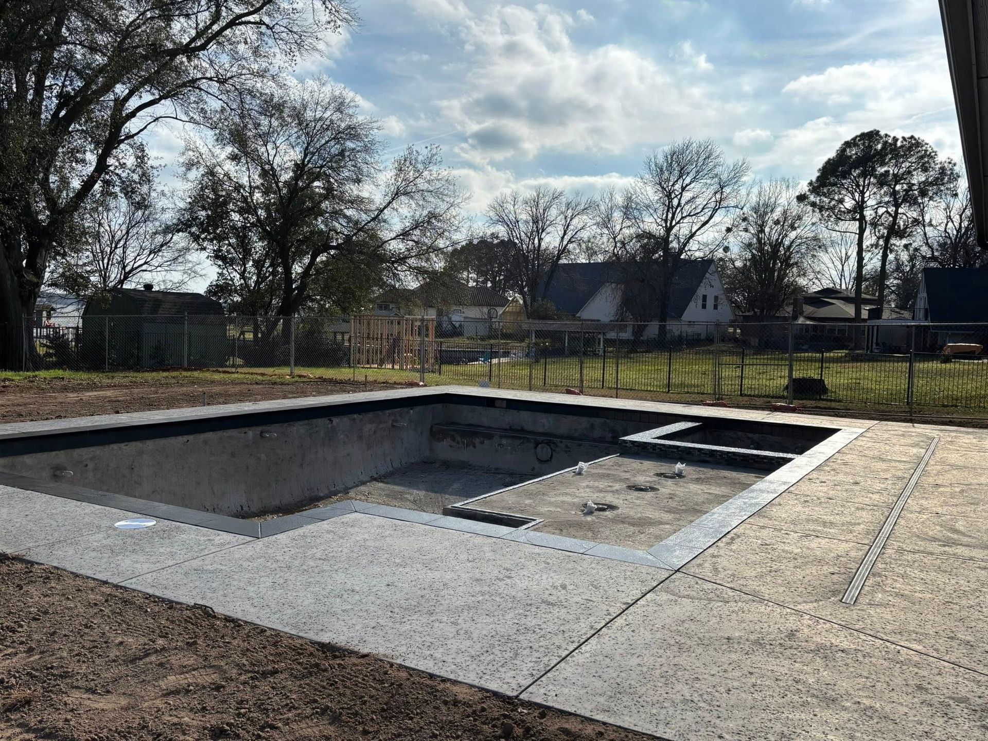 An empty, newly constructed concrete swimming pool with a surrounding stone patio set against a background of trees.