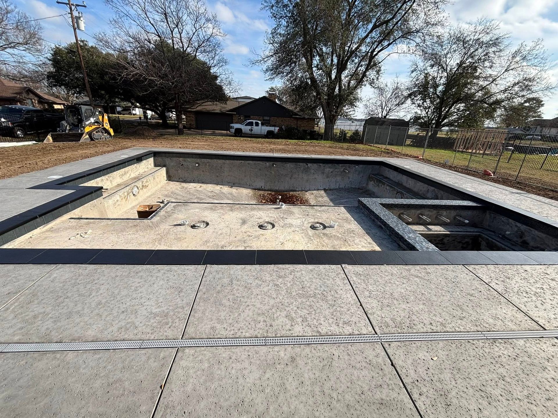 An empty swimming pool construction site featuring concrete decking, steps, and a hot tub area under a clear blue sky.