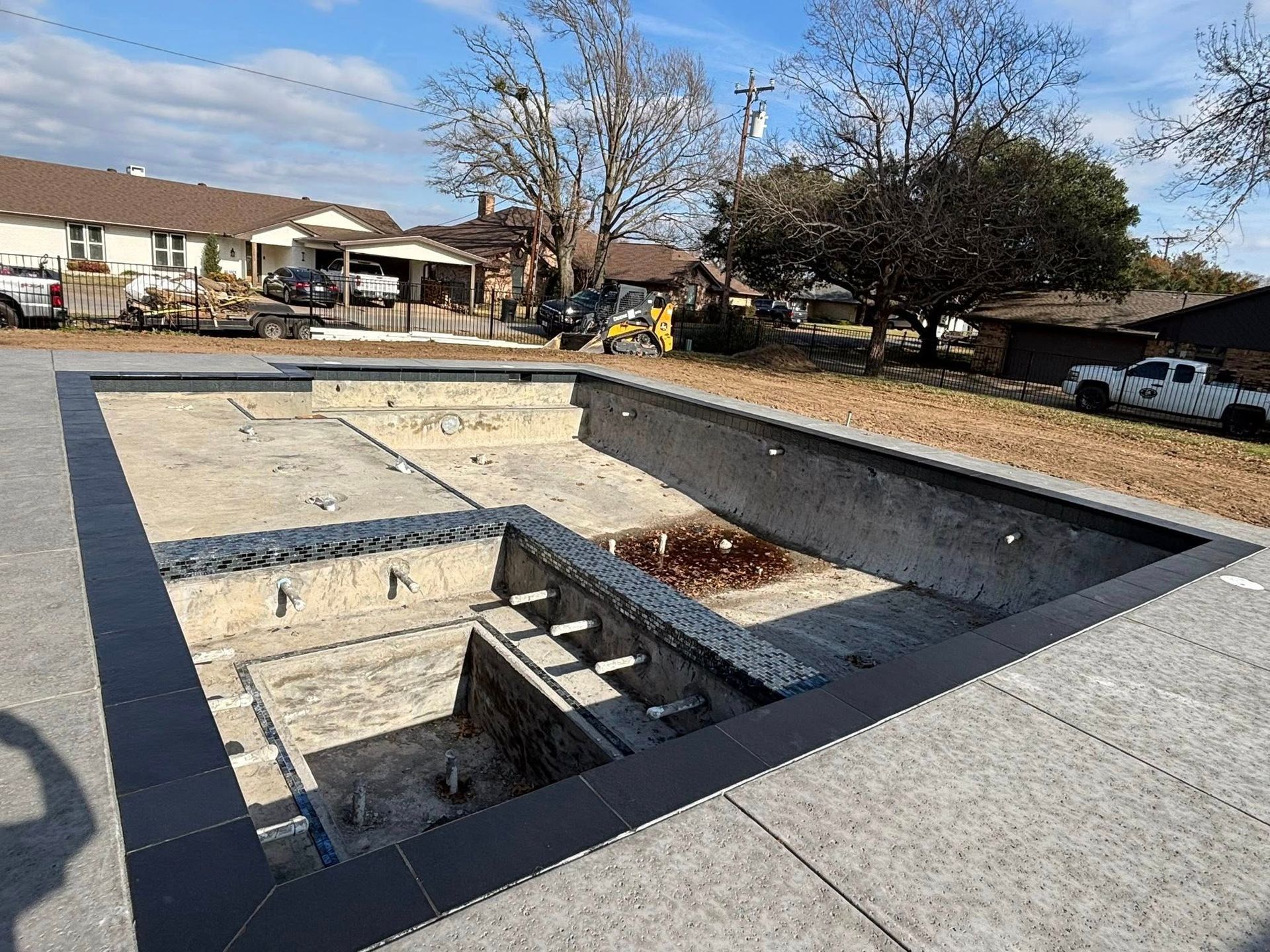 An unfinished, empty concrete swimming pool featuring a built-in spa area, surrounded by dark tiled coping and pavers.